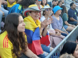 Scenes from the Columbia v. Portugal watch party at East Boston Stadium Spectators at a stadium, a woman with a striped hat and a Colombian flag draped over her shoulders, clapping.
