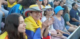 Scenes from the Columbia v. Portugal watch party at East Boston Stadium Spectators at a stadium, a woman with a striped hat and a Colombian flag draped over her shoulders, clapping.