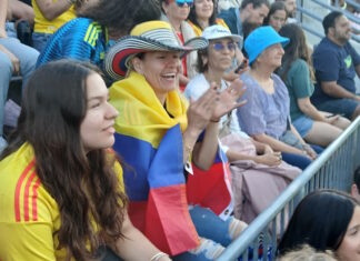 Scenes from the Columbia v. Portugal watch party at East Boston Stadium Spectators at a stadium, a woman with a striped hat and a Colombian flag draped over her shoulders, clapping.