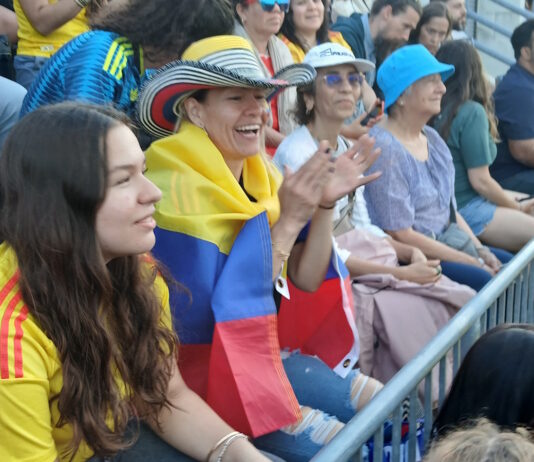 Scenes from the Columbia v. Portugal watch party at East Boston Stadium Spectators at a stadium, a woman with a striped hat and a Colombian flag draped over her shoulders, clapping.