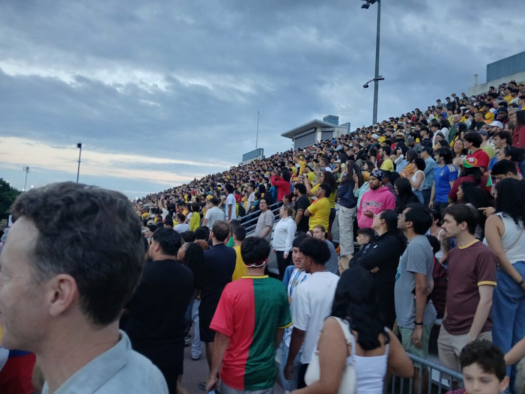 Crowded stadium bleachers filled with a diverse group of spectators watching an event at dusk/early evening.