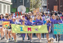 ZUMIX to host 2026 Walk for Music Diverse group marching in a street parade, holding a bright lime banner that reads ZUMIX and colorful signs around them.