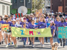 ZUMIX to host 2026 Walk for Music Diverse group marching in a street parade, holding a bright lime banner that reads ZUMIX and colorful signs around them.