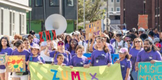 ZUMIX to host 2026 Walk for Music Diverse group marching in a street parade, holding a bright lime banner that reads ZUMIX and colorful signs around them.