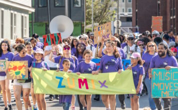 ZUMIX to host 2026 Walk for Music Diverse group marching in a street parade, holding a bright lime banner that reads ZUMIX and colorful signs around them.