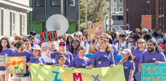 ZUMIX to host 2026 Walk for Music Diverse group marching in a street parade, holding a bright lime banner that reads ZUMIX and colorful signs around them.