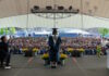 Excel Academy Charter High School celebrates Class of 2026 graduation Graduate wearing cap and gown speaks from a stage to a large outdoor audience under a white tent.