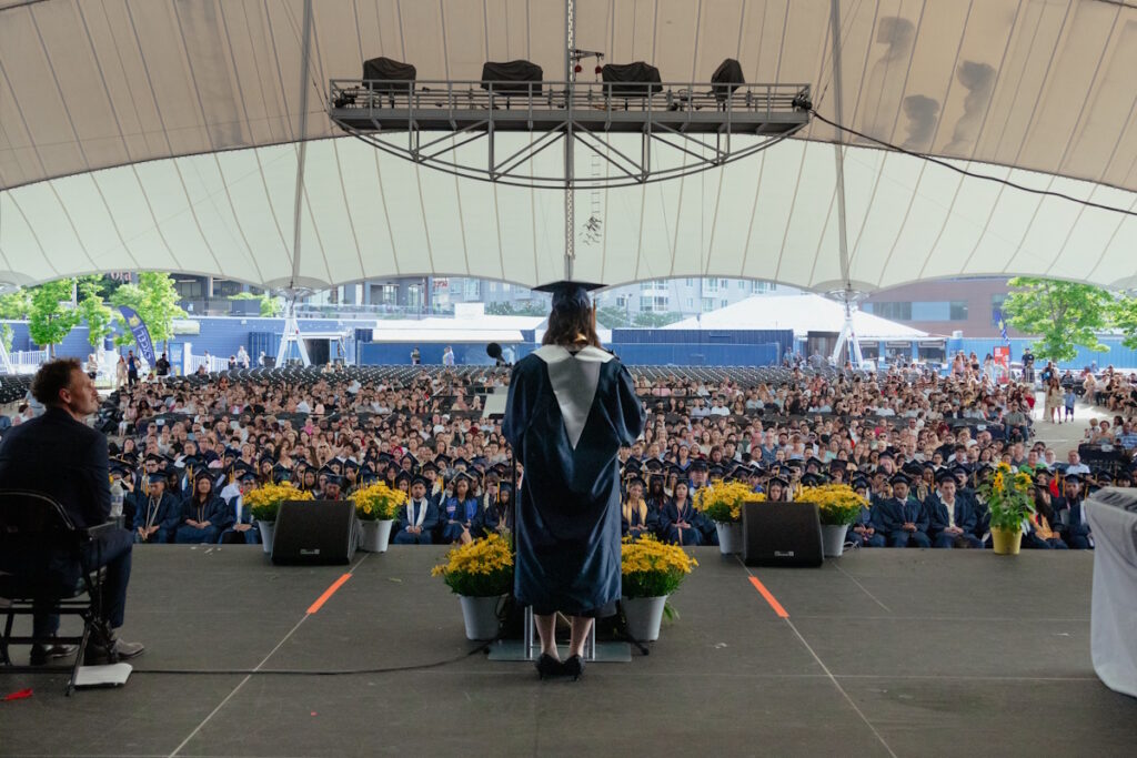 Graduate wearing cap and gown speaks from a stage to a large outdoor audience under a white tent.