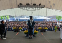 Excel Academy Charter High School celebrates Class of 2026 graduation Graduate wearing cap and gown speaks from a stage to a large outdoor audience under a white tent.