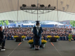 Excel Academy Charter High School celebrates Class of 2026 graduation Graduate wearing cap and gown speaks from a stage to a large outdoor audience under a white tent.