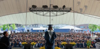 Excel Academy Charter High School celebrates Class of 2026 graduation Graduate wearing cap and gown speaks from a stage to a large outdoor audience under a white tent.