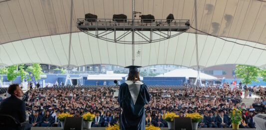 Excel Academy Charter High School celebrates Class of 2026 graduation Graduate wearing cap and gown speaks from a stage to a large outdoor audience under a white tent.
