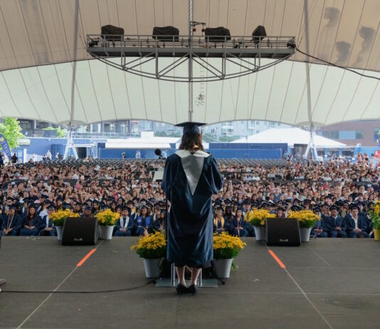 Excel Academy Charter High School celebrates Class of 2026 graduation Graduate wearing cap and gown speaks from a stage to a large outdoor audience under a white tent.