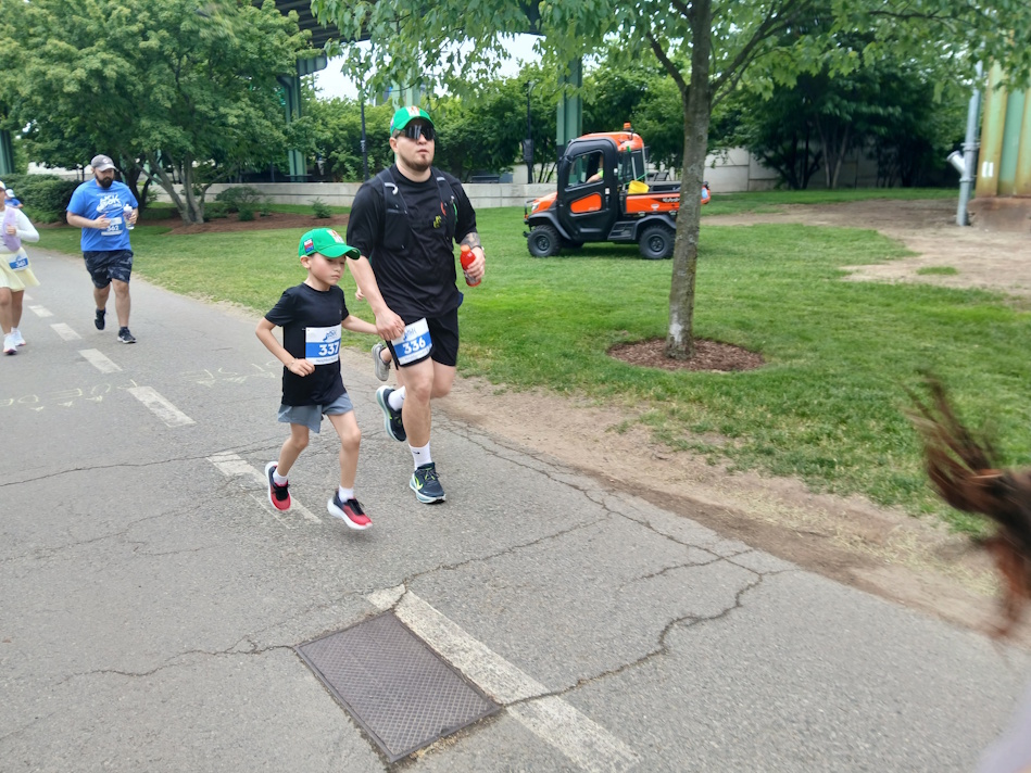 A man and young boy run together in a street race, wearing green caps and bib numbers, with spectators and a small orange vehicle in a park setting behind them.