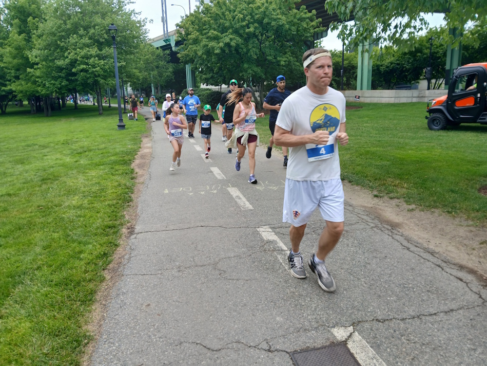Group of runners in a park race on a paved path, with a man in white shirt and bib number 4 leading the pack.