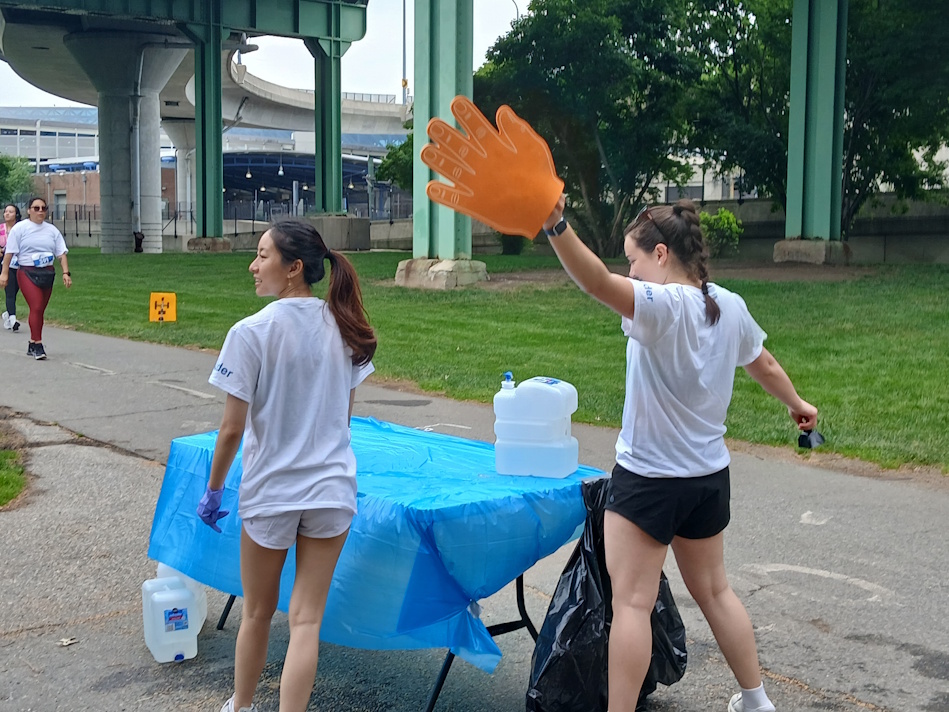 Two volunteers in white shirts stand by a blue-covered table distributing water at an outdoor event, one wearing an oversized orange glove as a playful cue.