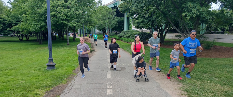Diverse group of runners and walkers on a paved park path; a child in a stroller is being pushed while others wear race bibs.