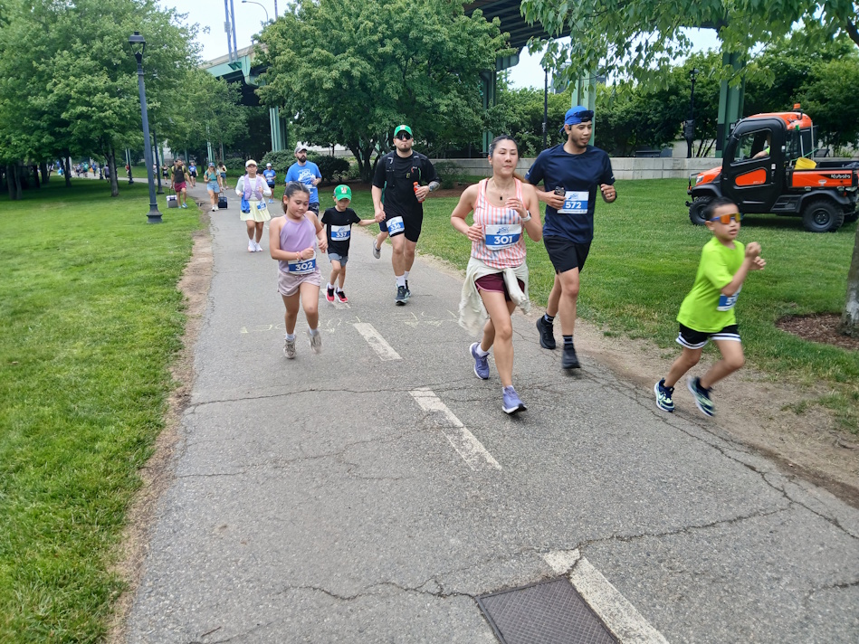 Group of runners, including children, sprinting on a park trail during a race with bib numbers visible on their shirts.