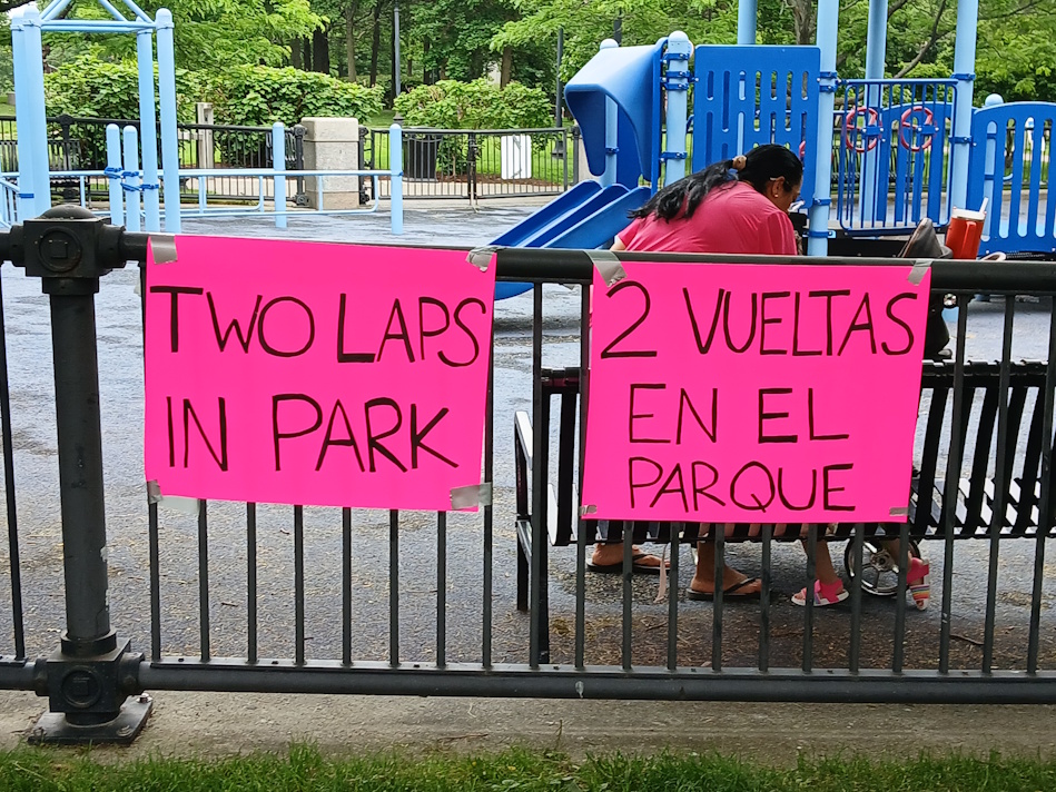 Bright pink sign on a black park fence reads 'Two laps in park' near a blue playground; a person in a pink shirt sits nearby.