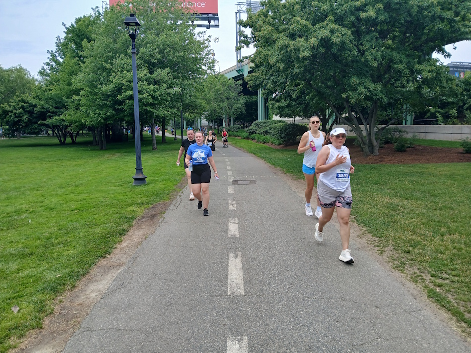 Runners in a park race on a paved path, bib numbers visible on their shirts.