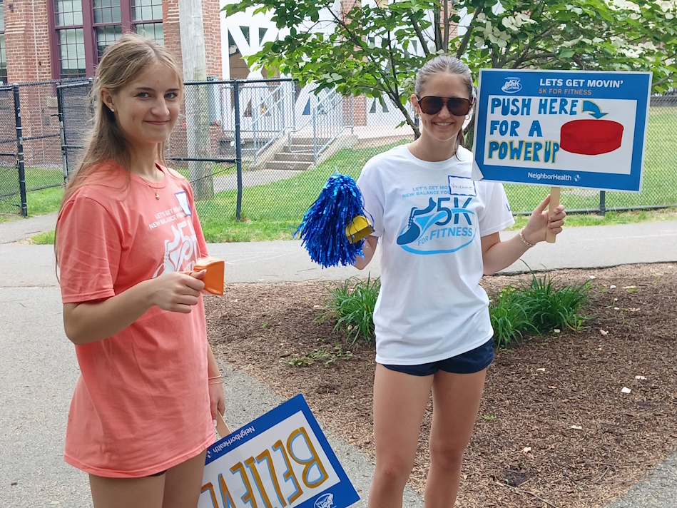 Two girls stand outside at a yard event; one in a coral T‑shirt holds an orange box, the other in a white tee with sunglasses holds a blue pom‑pom and a sign that says “Push Here For A Power Up.”