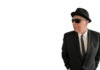 Sal Baglio’s new record is coming out in July Man in a black suit and tie wearing a hat and sunglasses, looking to the right with hands in pockets.