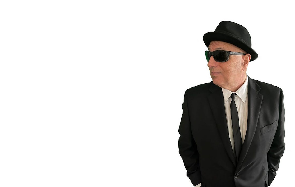 Man in a black suit and tie wearing a hat and sunglasses, looking to the right with hands in pockets.
