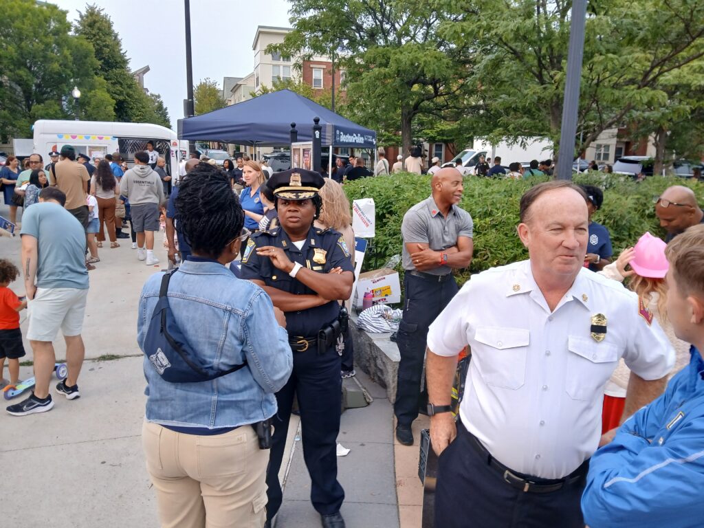 Crowd of people at an outdoor community event with police officers near a blue tent and trees in the background.