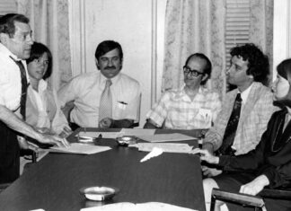 Gina Fiandaca: Appreciating John Vitagliano, who always believed in people Six professionals in a office meeting around a long conference table; a man stands presenting papers to seated colleagues.
