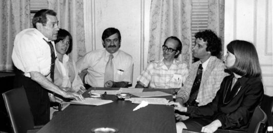 Gina Fiandaca: Appreciating John Vitagliano, who always believed in people Six professionals in a office meeting around a long conference table; a man stands presenting papers to seated colleagues.