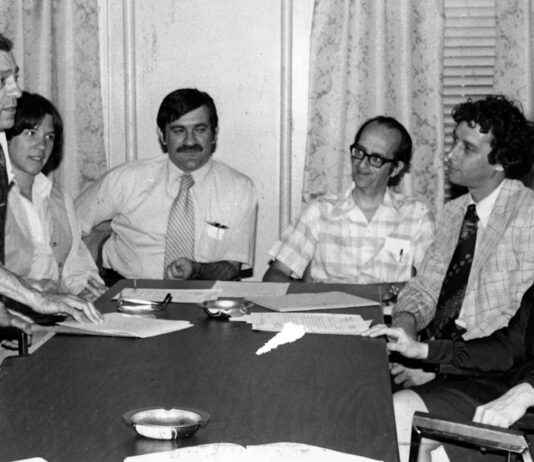 Gina Fiandaca: Appreciating John Vitagliano, who always believed in people Six professionals in a office meeting around a long conference table; a man stands presenting papers to seated colleagues.