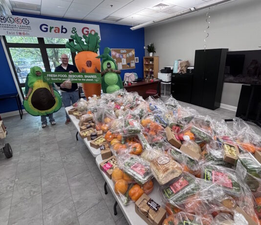 Maverick Landing Community Services launches Grab & Go produce market, expanding affordable fresh food access across East Boston Volunteers in avocado and carrot costumes pose behind tables stacked with grab-and-go produce bags at a community food drive.