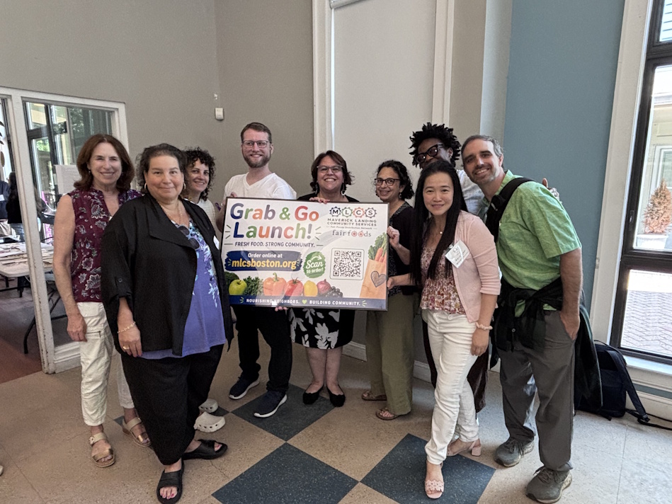 Nine diverse adults stand indoors, smiling and holding a colorful 'Grab & Go Launch' banner for a community food program.
