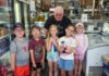 Learning how food brings us together Six children and an older man pose in a grocery/deli shop, standing behind a crate of red bell peppers on the floor.