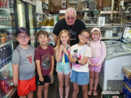 Learning how food brings us together Six children and an older man pose in a grocery/deli shop, standing behind a crate of red bell peppers on the floor.