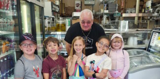 Learning how food brings us together Six children and an older man pose in a grocery/deli shop, standing behind a crate of red bell peppers on the floor.