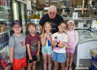 Learning how food brings us together Six children and an older man pose in a grocery/deli shop, standing behind a crate of red bell peppers on the floor.