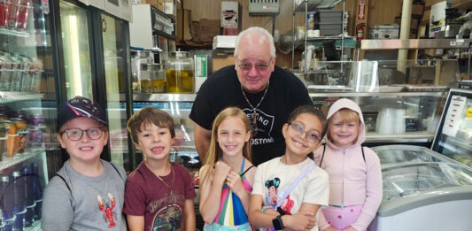Learning how food brings us together Six children and an older man pose in a grocery/deli shop, standing behind a crate of red bell peppers on the floor.