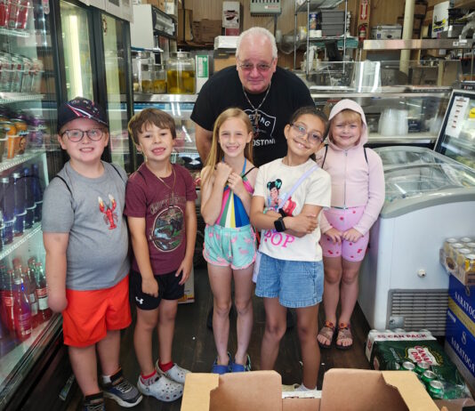 Learning how food brings us together Six children and an older man pose in a grocery/deli shop, standing behind a crate of red bell peppers on the floor.