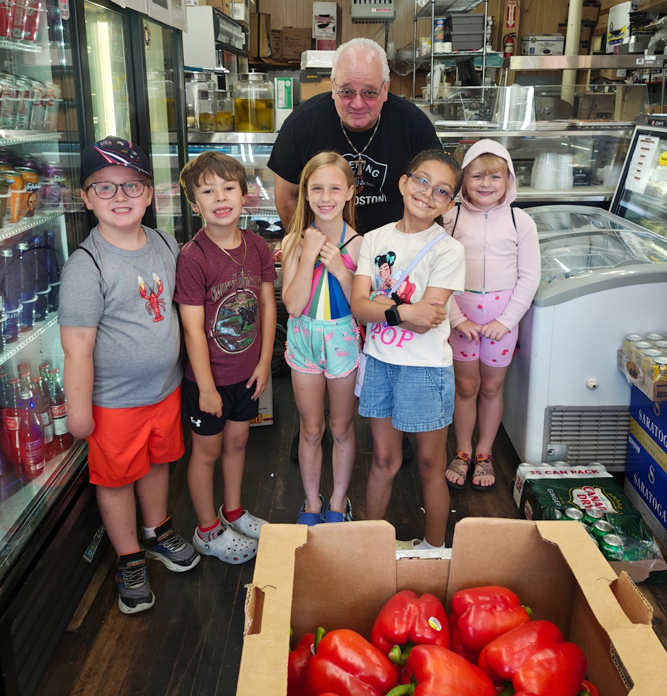 Six children and an older man pose in a grocery/deli shop, standing behind a crate of red bell peppers on the floor.