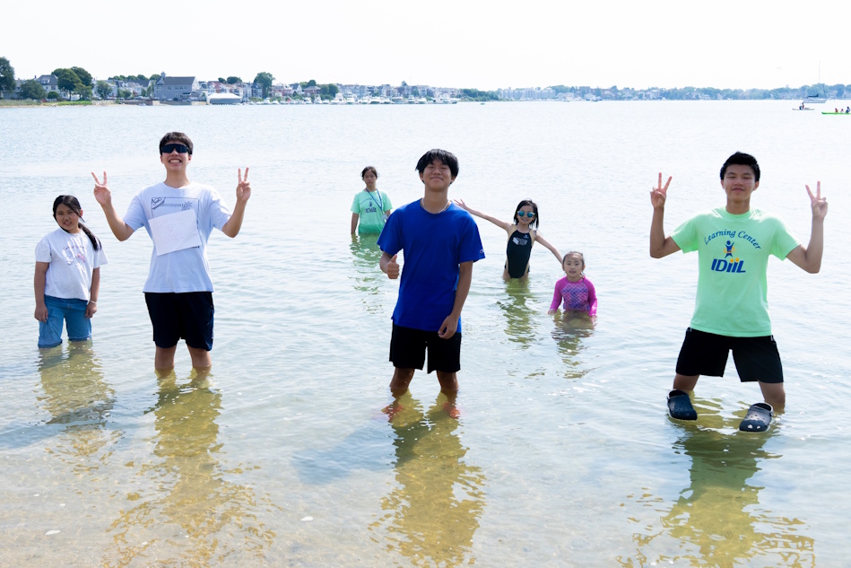 Group of children and teens standing in shallow beach water, some flashing peace signs with a coastal town in the distance.