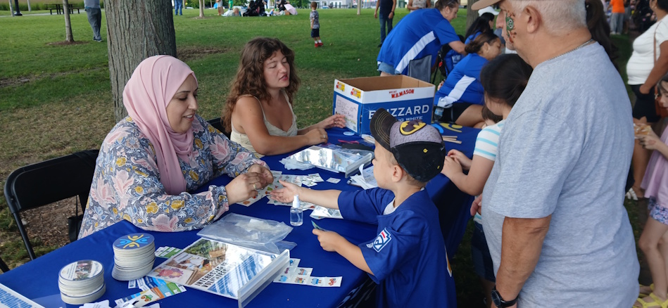 Woman in a pink hijab and floral blouse hands a small item to a young boy at a park community event table with blue cloth.