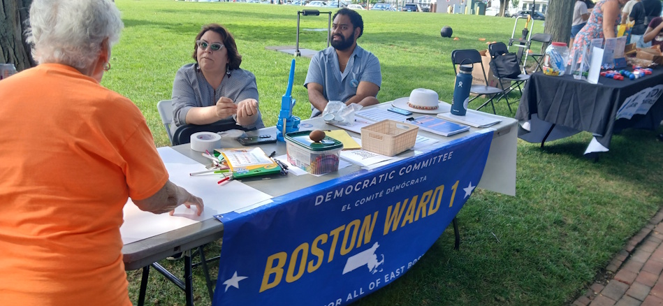 Community members sit at a park table for Boston Ward 1 Democratic Committee outreach, banner visible on front left.