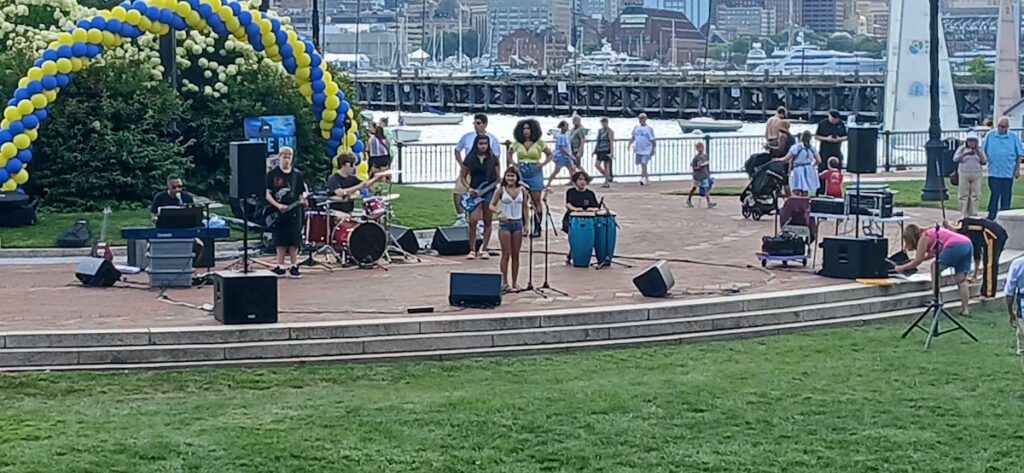 Live street band performing on a waterfront promenade under a yellow-and-blue balloon arch by the harbor.