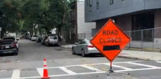 Residents and businesses are fed up with the parking situation in East Boston Orange 'Road Closed' sign with a black left arrow and a traffic cone on a city street near a crosswalk, indicating road construction.
