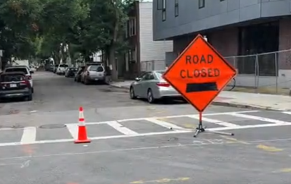 Residents and businesses are fed up with the parking situation in East Boston Orange 'Road Closed' sign with a black left arrow and a traffic cone on a city street near a crosswalk, indicating road construction.