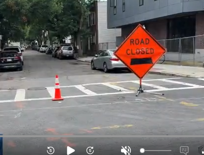 Orange 'Road Closed' sign with a black left arrow and a traffic cone on a city street near a crosswalk, indicating road construction.