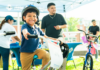 NeighborHealth promotes safe summer riding through its Annual Childrenโs Bike Safety Day Smiling young boy wearing a helmet rides a white bike at an outdoor event, others in background under a canopy