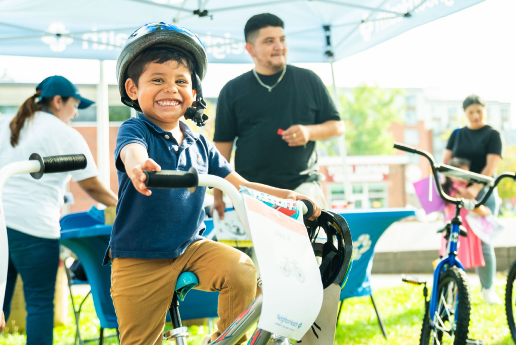 Smiling young boy wearing a helmet rides a white bike at an outdoor event, others in background under a canopy
