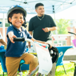 NeighborHealth promotes safe summer riding through its Annual Childrenโs Bike Safety Day Smiling young boy wearing a helmet rides a white bike at an outdoor event, others in background under a canopy